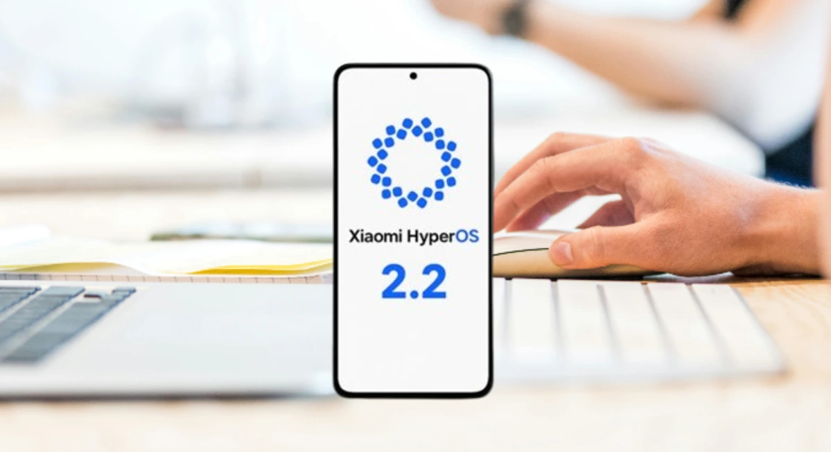An image shows a smartphone with the Xiaomi HyperOS logo and the number "2.2" highlighted on the screen. In the background, out of focus, a person uses a computer with a mouse and keyboard.