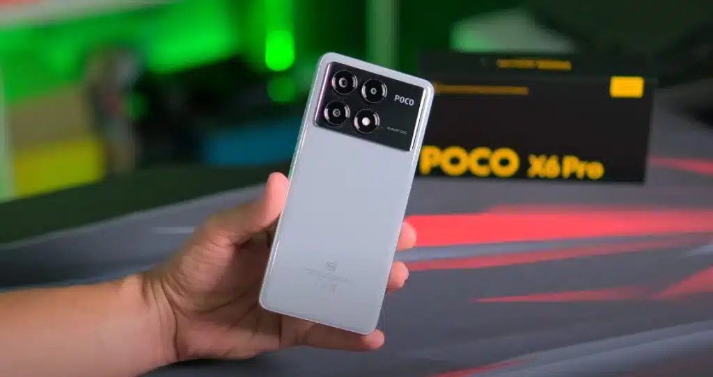 Hand holding the back of a gray/silver POCO X6 Pro smartphone, highlighting the black camera module and the "POCO" logo, with the device's box partially visible and a blurred background with green and red lights.
