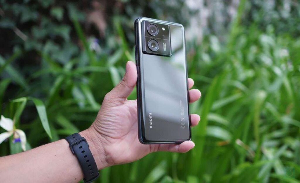 A hand holding a black Xiaomi 13T smartphone, showing the rear with the camera module and the Xiaomi logo, updated with HyperOS 2.2.
