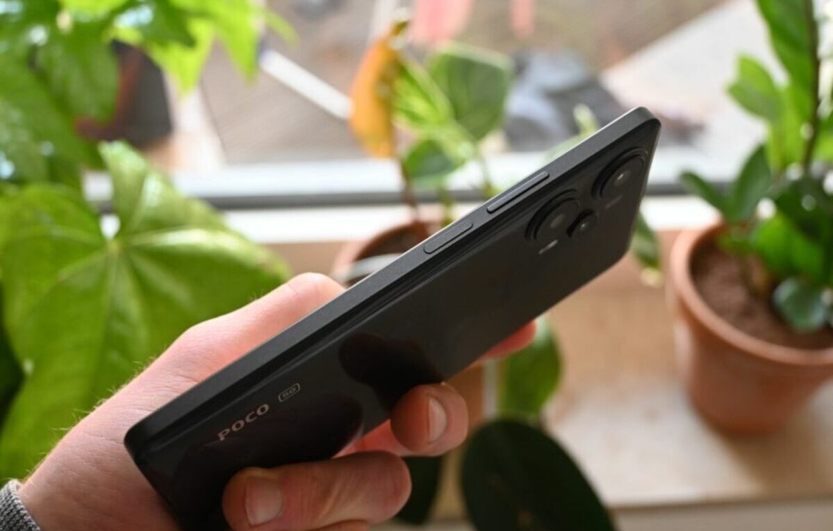 Hand holding a POCO F5, with a blurred background of plants. The device runs the POCO F5: HyperOS 2.2 Global Stable operating system.