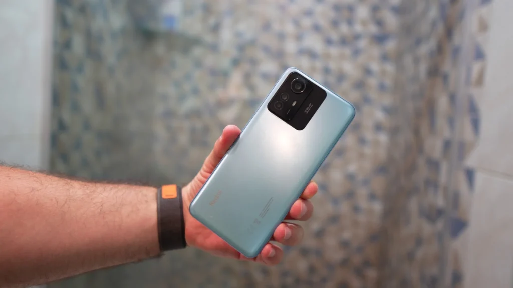 A hand holds a light blue Redmi Note 12S smartphone, showing the back of the device. The rear camera is prominent, with a large module that includes the 108MP main lens and other smaller lenses. The "Redmi" logo and some regulatory information are printed on the bottom of the device. The blurred background is a blue and white wall tile.
