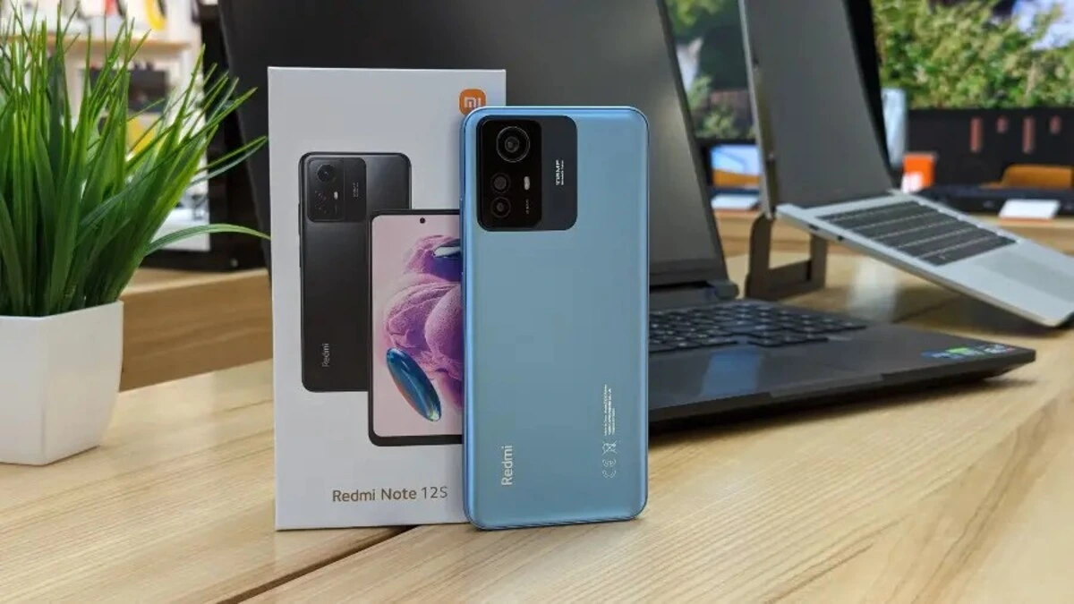 A light blue Redmi Note 12S smartphone sits vertically on a wooden table, with its back facing the camera, displaying the camera module design and the "Redmi" branding. Next to it, the white Redmi Note 12S box features an image of the phone in black. In the background, in the background, a black laptop, a silver laptop and a potted green plant can be seen in the background, suggesting a tech review environment. This device is eligible for the HyperOS 2.2 update.