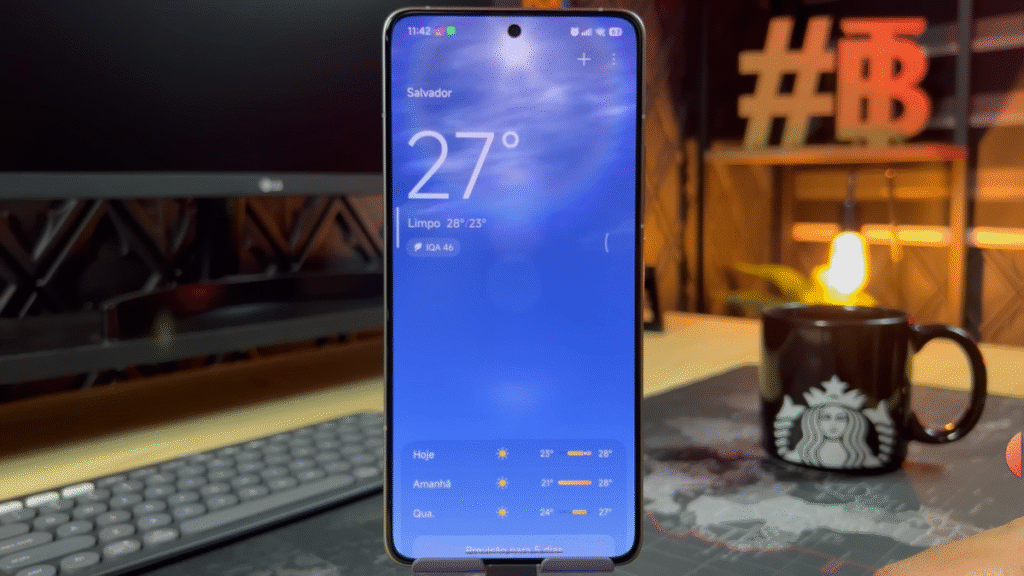 Xiaomi smartphone with the HyperOS 3 Weather app displaying the temperature of 27 degrees in Salvador, Brazil, and the forecast for the next few days, on a table setup with a monitor and decorative objects.