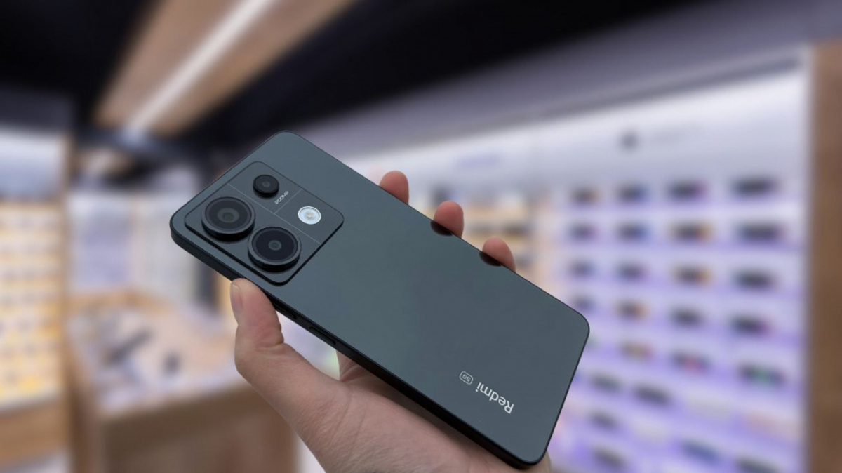 A dark Redmi smartphone being held in a person's hand, with its triple camera module and flash visible on the back.