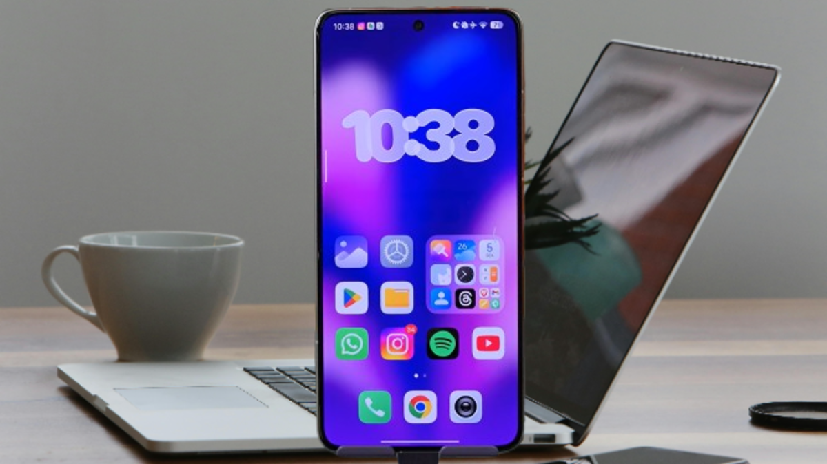A smartphone on a work desk displays the HyperOS Launcher 3 home screen with a purple-blue wallpaper, a clock widget, and app icons, next to a silver laptop and a cup of coffee.