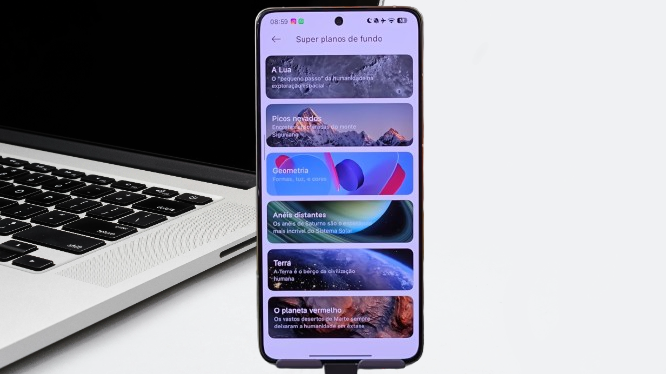 Xiaomi smartphone displaying HyperOS 3's "Super Wallpapers" screen, with options such as The Moon, Snowy Peaks, Geometry, Distant Rings, Earth, and The Red Planet, and a laptop in the background.