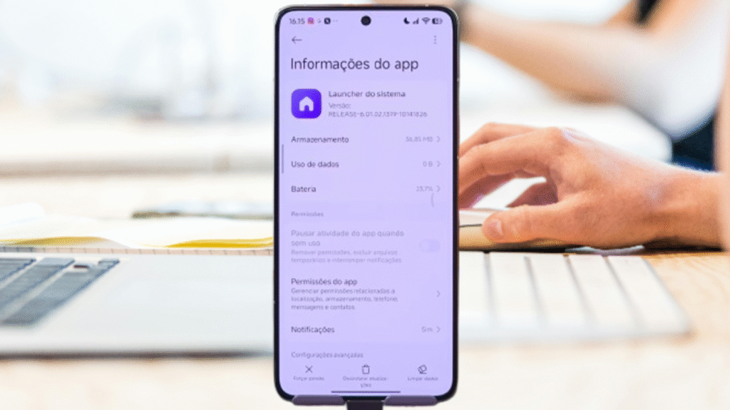 The image shows a modern smartphone with thin bezels, focused on the screen displaying the "App Info" interface for the "System Launcher," which is part of HyperOS Launcher 3. The screen's background is a light lilac. At the top, the phone's status bar is visible, followed by the "App Info" title and a back arrow to the left.The interface shows the purple "System Launcher" app icon and the version "RELEASE-6.01.32.1379-1018120." Below, the "Storage," "Data Usage," and "Battery" sections are listed with their respective information. Further down, the "Permissions" section details options such as "Pause app activity when idle," "App Permissions," and "Notifications." At the bottom of the screen, there are options to "Force Stop," "Uninstall Updates," and "Clear Data."The smartphone is positioned vertically on a stand on a light wood table. In the blurred background, elements of a work environment are visible, including part of a silver laptop on the left and a white keyboard with a person's hand operating a mouse on the right. The image is well-lit and clear, highlighting the details of the HyperOS Launcher 3 user interface.