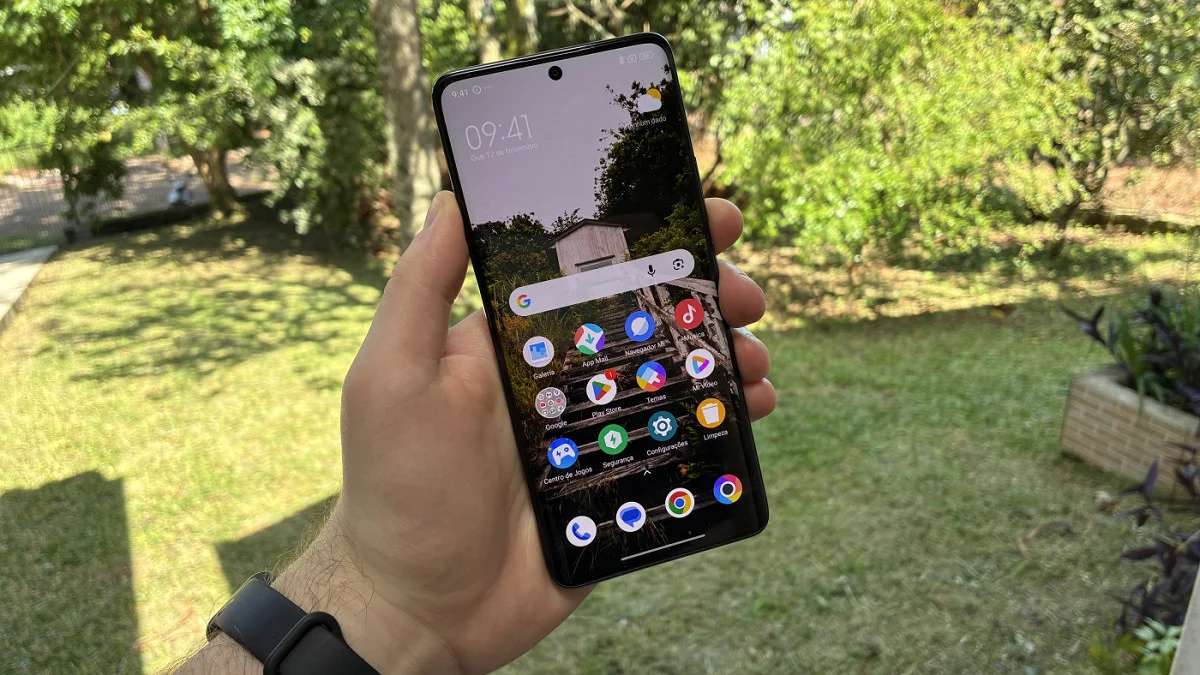 A person holding a Xiaomi smartphone outdoors showing the HyperOS 3 Global home screen interface with a nature wallpaper and various app icons.